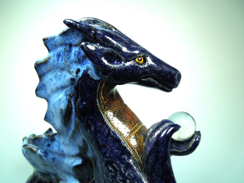 May include: A ceramic dragon figurine in shades of blue and brown, holding a clear glass orb. The dragon has a detailed, textured surface with yellow eyes. The figurine is a decorative item.