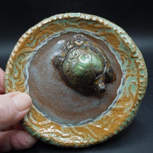 May include: A ceramic turtle figurine on a round, decorative plate. The turtle is green and brown, centered on a brown base. The plate has a detailed, textured rim with shades of green and brown.