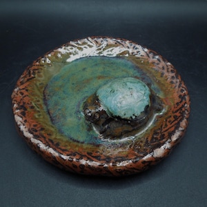 May include: A handcrafted ceramic dish with a turtle in a pond design. The dish has a brown, green, and blue colour scheme. The turtle is a light blue colour, and the pond is a mix of green and blue. The dish is round and has a textured rim.