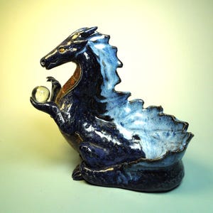 May include: A ceramic dragon sculpture in shades of blue, with a lighter blue and white mane and a small, clear glass orb held in its hand. The dragon has a small bowl-shaped opening on its back.