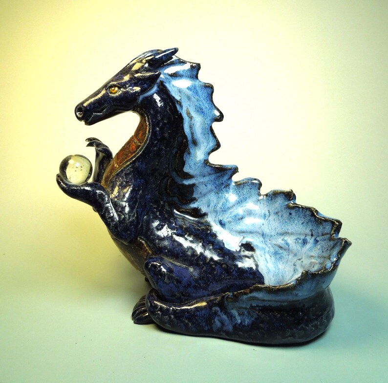 May include: A ceramic dragon sculpture in shades of blue, with a lighter blue and white mane and a small, clear glass orb held in its hand. The dragon has a small bowl-shaped opening on its back.
