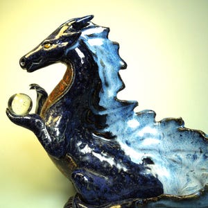 May include: A ceramic dragon sculpture in shades of blue, with a lighter blue and white mane. The dragon holds a clear glass orb in its claw. The sculpture has a detailed, textured surface and a touch of gold accents.