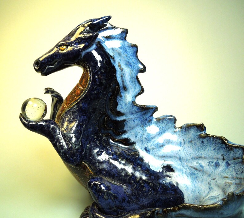 May include: A ceramic dragon sculpture in shades of blue, with a lighter blue and white mane. The dragon holds a clear glass orb in its claw. The sculpture has a detailed, textured surface and a touch of gold accents.