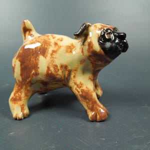 Puede incluir: Una figura de carlino de cer&aacute;mica con acabado brillante, cuerpo color crema con manchas marrones. El perro tiene una cara negra distintiva, una cola corta erguida y cuatro patas robustas, representado en posici&oacute;n de pie con la cabeza inclinada.