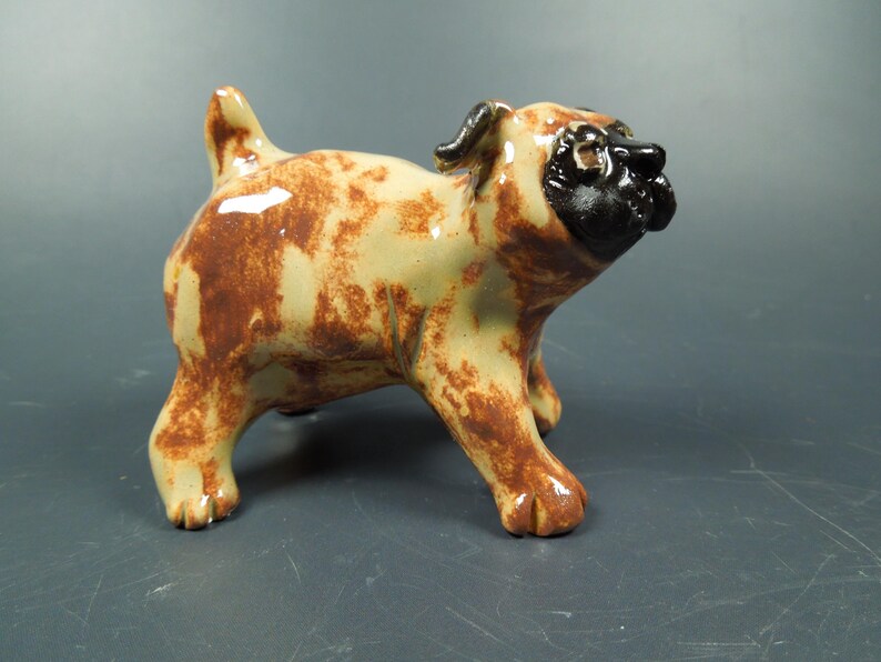Puede incluir: Una figura de carlino de cer&aacute;mica con acabado brillante, cuerpo color crema con manchas marrones. El perro tiene una cara negra distintiva, una cola corta erguida y cuatro patas robustas, representado en posici&oacute;n de pie con la cabeza inclinada.