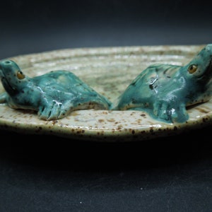 May include: A ceramic dish with a speckled glaze features two small, teal-colored turtle figurines. The turtles are facing each other, with their heads raised.
