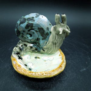 May include: A ceramic snail figurine with a blue and black speckled shell and a light green body. The snail sits on a small, shallow dish with a gold-colored rim. The figurine is a decorative item.