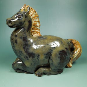 May include: A ceramic horse figurine in a resting pose. The horse has a dark grey body with brown and tan accents on the mane and tail. The figurine is glazed and has a glossy finish.