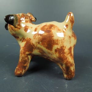 Puede incluir: Una peque&ntilde;a figura de perro de cer&aacute;mica con acabado brillante, que presenta una base de color crema con manchas marrones irregulares. El perro tiene orejas de color marr&oacute;n oscuro y una cola corta y erguida, vista desde atr&aacute;s.