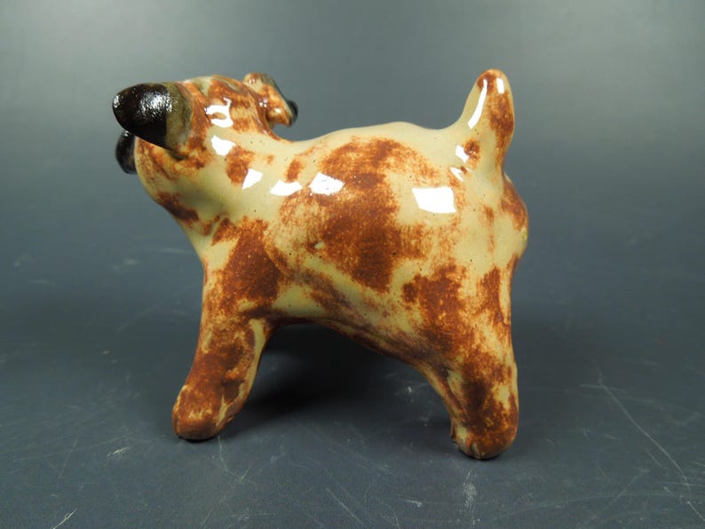 Puede incluir: Una peque&ntilde;a figura de perro de cer&aacute;mica con acabado brillante, que presenta una base de color crema con manchas marrones irregulares. El perro tiene orejas de color marr&oacute;n oscuro y una cola corta y erguida, vista desde atr&aacute;s.