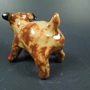 Puede incluir: Una peque&ntilde;a figura de cer&aacute;mica de un perro con un acabado brillante. El perro tiene un pelaje base de color crema con manchas marrones irregulares que cubren su cuerpo, patas y cola. Las orejas son de color marr&oacute;n oscuro.