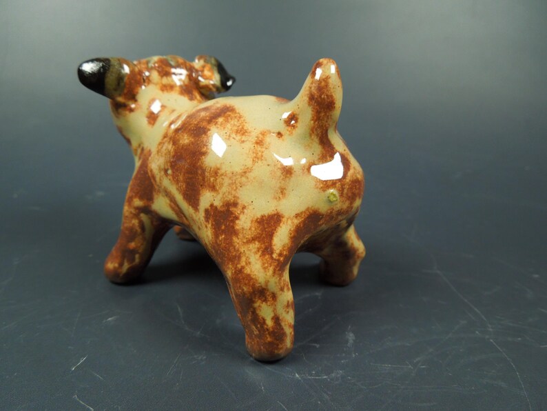 Puede incluir: Una peque&ntilde;a figura de cer&aacute;mica de un perro con un acabado brillante. El perro tiene un pelaje base de color crema con manchas marrones irregulares que cubren su cuerpo, patas y cola. Las orejas son de color marr&oacute;n oscuro.