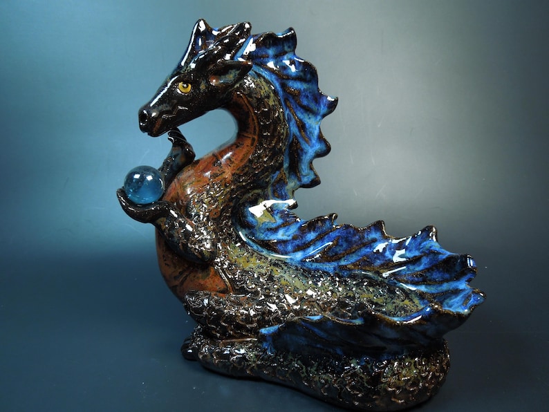 K&ouml;nnte beinhalten: Keramikdrachenskulptur in Braun-, Schwarz- und Blaut&ouml;nen. Der Drache h&auml;lt eine blaue Glaskugel. Die Skulptur hat eine strukturierte Oberfl&auml;che und ist mit erhobenem Kopf dargestellt. Die Fl&uuml;gel und der R&uuml;cken des Drachen sind leuchtend blau.
