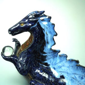 May include: A ceramic dragon sculpture in shades of blue, with a clear glass orb held in its clawed hand. The dragon has a textured, scaled appearance, with a lighter blue mane and accents of brown and gold.