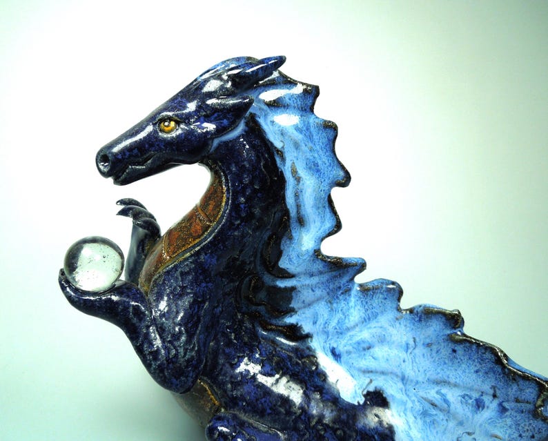 May include: A ceramic dragon sculpture in shades of blue, with a clear glass orb held in its clawed hand. The dragon has a textured, scaled appearance, with a lighter blue mane and accents of brown and gold.