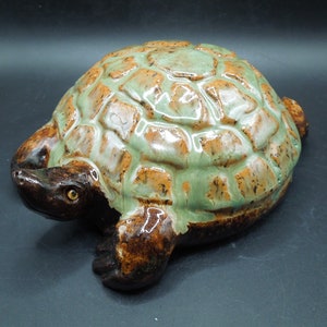 May include: A ceramic turtle ornament with a glossy finish. The shell is a mosaic of green and brown, while the head and limbs are dark brown. The eyes are bright yellow. The ornament is a decorative item.