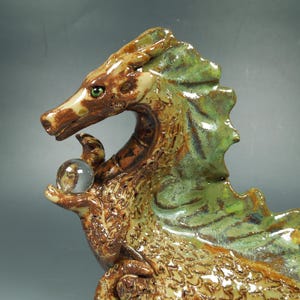 May include: A ceramic dragon sculpture featuring a glossy glaze in shades of brown and green. The dragon has a textured, scaled body, a prominent dorsal fin, and a single green eye. It holds a small, clear, spherical glass orb in its claw.