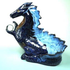 May include: A ceramic dragon figurine in shades of blue, with a clear glass orb held in its claw. The dragon has a textured, scaled appearance and a detailed, expressive face. The figurine is approximately 10 inches tall.