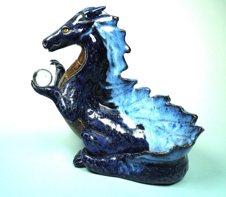 May include: A ceramic dragon figurine in shades of blue, with a clear glass orb held in its claw. The dragon has a textured, scaled appearance and a detailed, expressive face. The figurine is approximately 10 inches tall.