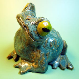 May include: A ceramic frog figurine with a textured blue and brown glaze. The frog has one large, prominent yellow eye with a black pupil, a wide mouth, and detailed webbed feet. The surface of the sculpture features a mottled finish.