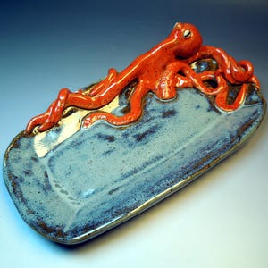 May include: A rectangular ceramic tray with a glossy blue and grey mottled glaze. A sculpted, bright orange octopus with visible suction cups on its tentacles is attached to the top edge, with its arms draped across the surface of the tray.