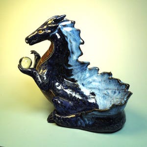 May include: A ceramic dragon sculpture in shades of blue, with a white and brown accent. The dragon holds a clear glass orb in its clawed hand. The sculpture has a bowl-like opening on its back.