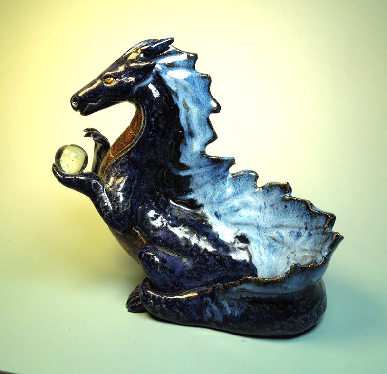 May include: A ceramic dragon sculpture in shades of blue, with a white and brown accent. The dragon holds a clear glass orb in its clawed hand. The sculpture has a bowl-like opening on its back.