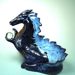 May include: A ceramic dragon sculpture in shades of blue, with a clear glass orb held in its claw. The dragon has a textured, glossy finish, with a lighter blue on its back and a darker blue on its body. The sculpture is a decorative item.