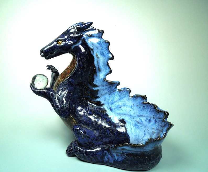 May include: A ceramic dragon sculpture in shades of blue, with a clear glass orb held in its claw. The dragon has a textured, glossy finish, with a lighter blue on its back and a darker blue on its body. The sculpture is a decorative item.