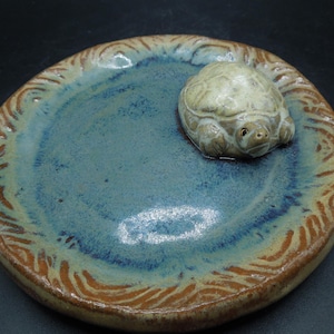 May include: A handmade ceramic dish with a turtle figurine. The dish has a blue interior with brown and tan accents around the rim. The turtle is light green and tan. The dish is round and has a rustic, artisanal feel.