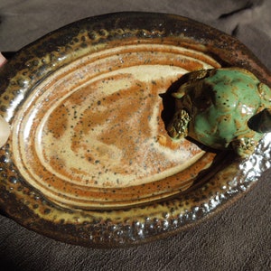 May include: A small, oval-shaped ceramic dish with a brown and tan glaze. The dish has a raised rim and a textured surface. A small green ceramic turtle figurine is sitting on the dish.