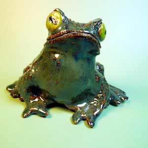 May include: A ceramic frog figurine with a glossy dark blue-green glaze and textured brown spots. The frog has large, prominent, light green eyes with dark pupils, a wide mouth, and four webbed feet positioned in a seated, upright posture.