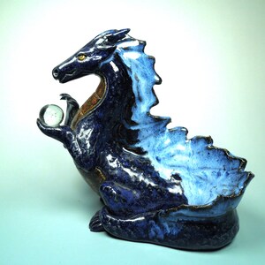 May include: A ceramic dragon sculpture in shades of blue, with a clear glass orb held in its clawed hand. The dragon has a textured, glossy finish and a detailed, stylized design. The sculpture is approximately 10 inches long.