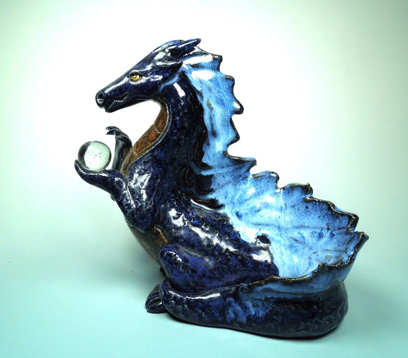 May include: A ceramic dragon sculpture in shades of blue, with a clear glass orb held in its clawed hand. The dragon has a textured, glossy finish and a detailed, stylized design. The sculpture is approximately 10 inches long.