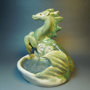 May include: A ceramic sculpture of a mythical sea creature with a horse-like head and a fish tail, featuring a glossy light green glaze with darker green accents. The creature is positioned behind a shallow, bowl-shaped base.