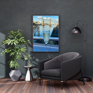 May include: A framed photograph of the Tower Bridge in London, with a glass of champagne in the foreground. The image is displayed in a living room setting, with a black armchair, a floor lamp, and a potted plant.