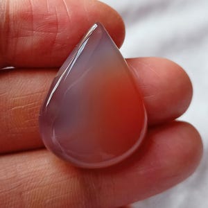 Op de afbeelding: Een druppelvormige edelsteen met een gepolijst oppervlak. De steen heeft een kleurverloop van licht lavendel naar oranjerood. De edelsteen wordt in een hand gehouden.