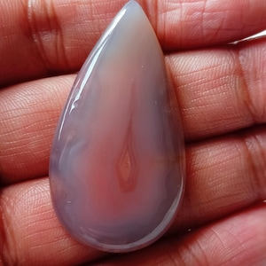 Könnte beinhalten: Ein polierter, tropfenförmiger Edelstein mit einer Mischung aus Farben. Der Stein weist einen Farbverlauf aus sanften Farbtönen auf, darunter Lavendel, Rosa und Weiß, mit einer durchscheinenden Qualität. Der Edelstein wird in einer Hand gehalten.