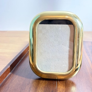 May include: A polished gold-tone metal picture frame with a rounded rectangular shape and a smooth, reflective surface. The frame features a thick, curved border surrounding a central rectangular opening that contains a plain, light beige insert.