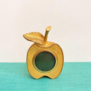 May include: A gold-toned metal apple-shaped picture frame with a textured finish and a single leaf extending from the top stem. The circular center opening is framed by a smooth gold border, designed to hold a small photograph.