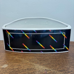 May include: A semi-circular ceramic wall planter with a glossy black exterior featuring a white rectangular border and diagonal rainbow-colored stripes. The interior is white and includes two small circular holes for mounting on a wall surface.