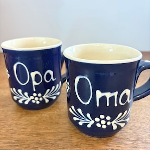 Puede incluir: Dos tazas de cerámica azul oscuro con interior color crema. Cada taza presenta texto blanco pintado a mano, una dice 'Opa' y la otra 'Oma'. Ambas tazas están decoradas con un patrón floral blanco en la base y a los lados del texto.