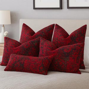 Puede incluir: Una colección de cojines rojos con un patrón oscuro y texturizado. Los cojines están colocados sobre una cama, mostrando su diseño decorativo. Los cojines varían en tamaño y forma, ofreciendo una estética acogedora y elegante para la decoración del hogar.
