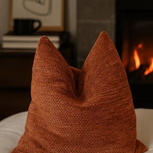 Könnte beinhalten: Ein strukturiertes, rostfarbenes Zierkissen mit spitzer Spitze. Das Kissen liegt auf einer weißen Oberfläche, mit einem Kamin und Kunstwerken im Hintergrund. Der Stoff des Kissens hat ein gewebtes Aussehen.
