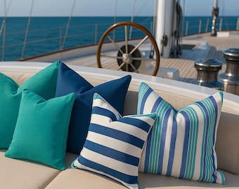 Könnte beinhalten: Eine Ansammlung von Zierkissen in Türkis, Blau und Weiß, angeordnet auf einem cremefarbenen Outdoor-Sofa. Einige Kissen sind einfarbig, andere haben Streifenmuster. Der Hintergrund zeigt ein Yachtdeck und das Meer.