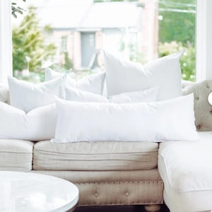 May include: A collection of white pillows of various sizes arranged on a beige sofa. The pillows include square, rectangular, and long bolster shapes. The sofa is positioned in front of a large window with a view of greenery.