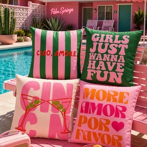 May include: A selection of decorative cushions featuring bold text and graphics. Designs include striped patterns, cocktail glasses, and phrases like "Girls Just Wanna Have Fun" and "More Amor Por Favor." The cushions are in shades of pink and green.