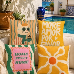 Pode incluir: Quatro almofadas decorativas com designs coloridos. Uma apresenta um gato com óculos de sol e cachecol, segurando uma xícara. Outra diz "MORE AMOR POR FAVOR". As outras duas almofadas têm um design floral e "HOME SWEET HOME".