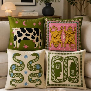 Puede incluir: Cuatro cojines cuadrados con diseños gráficos distintos: una vaca blanca y negra sobre verde, dos leopardos amarillos sobre rosa, dos serpientes verdes sobre crema y dos rostros estilizados en verde sobre crema, con motivos celestiales y florales.