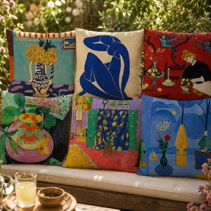 Puede incluir: Una colección de cojines decorativos con diseños artísticos y coloridos. Los cojines muestran varias pinturas con colores y patrones vibrantes, incluyendo arreglos florales, figuras y escenas de interiores. Los cojines están colocados en un banco blanco al aire libre.